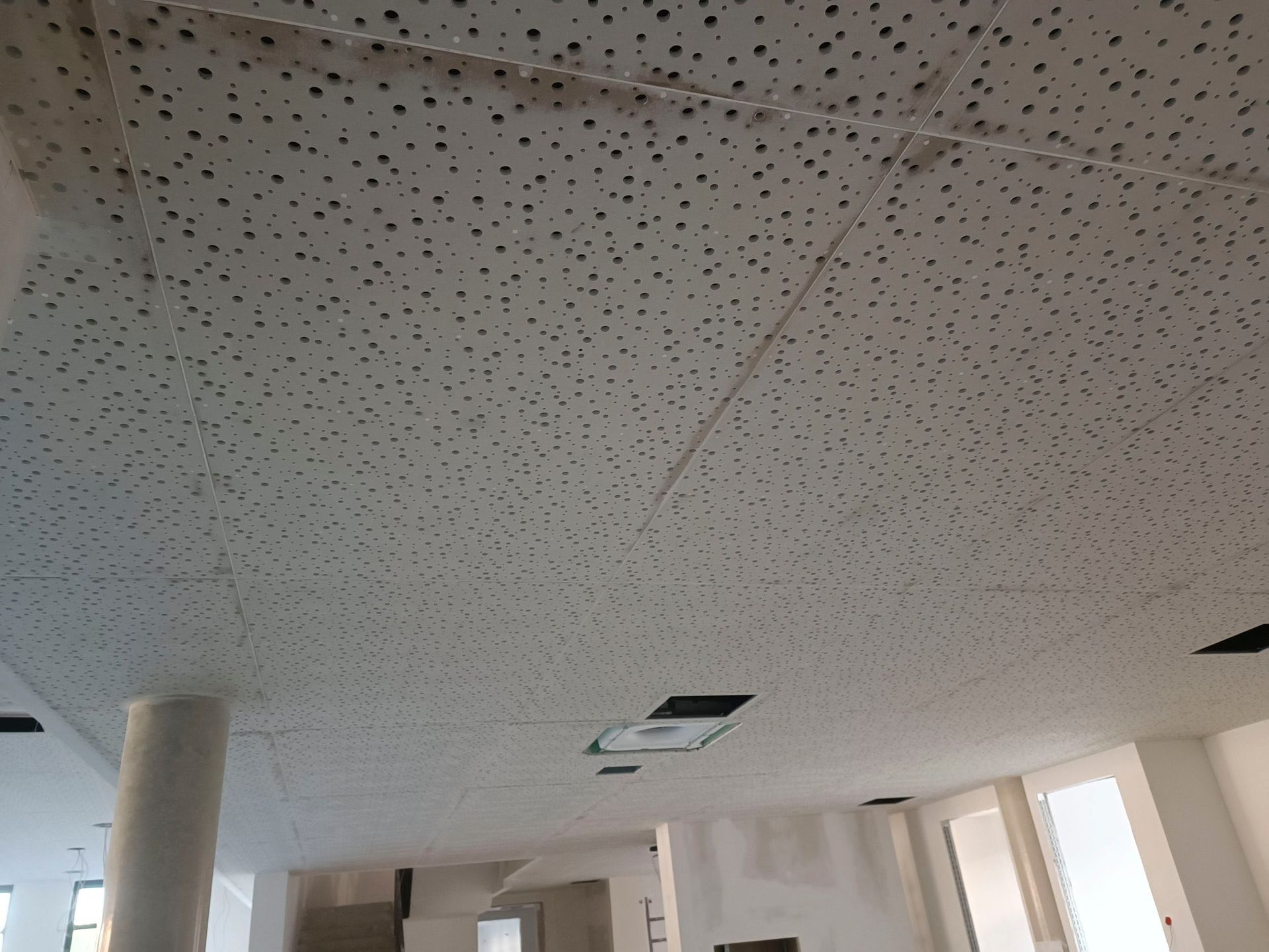 Weiße Akustikplattendecke mit kleinen schwarzen Perforationen. Einige Deckenleuchten sind sichtbar.