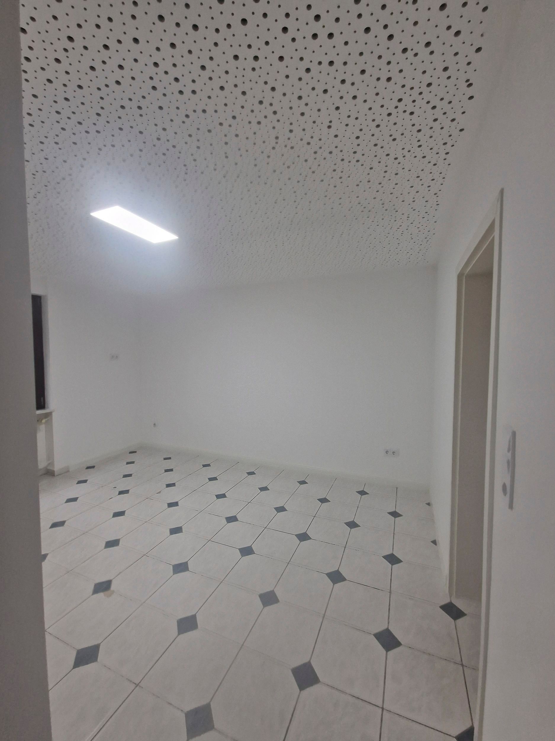 Leerer Raum mit weißen Wänden, gefliestem Boden und gesprenkelter Decke; eine Tür und ein Fenster sind sichtbar.