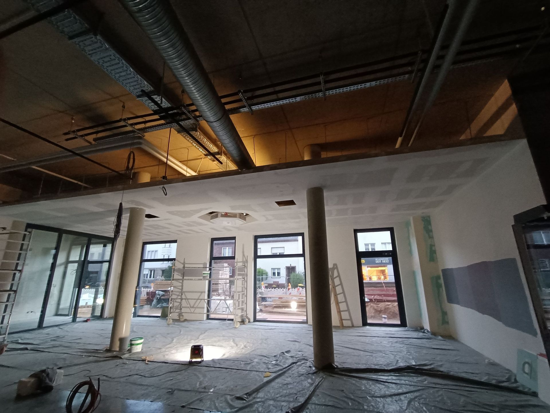 Innenansicht eines im Bau befindlichen Gebäudes mit unfertiger Decke, Stützsäulen und Fenstern.