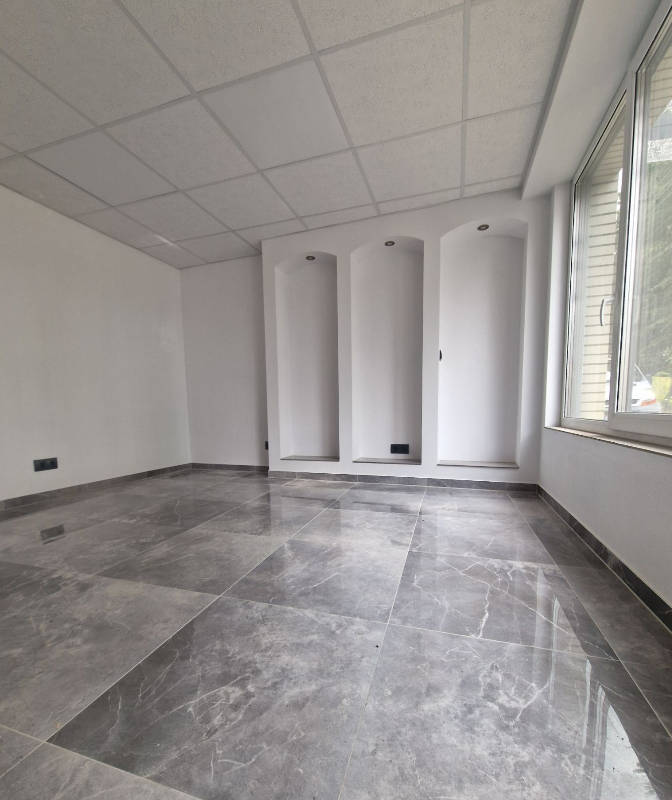 Leerer Raum mit grau gefliestem Boden, weißen Wänden, zurückgesetzten Wandnischen und einem Fenster.