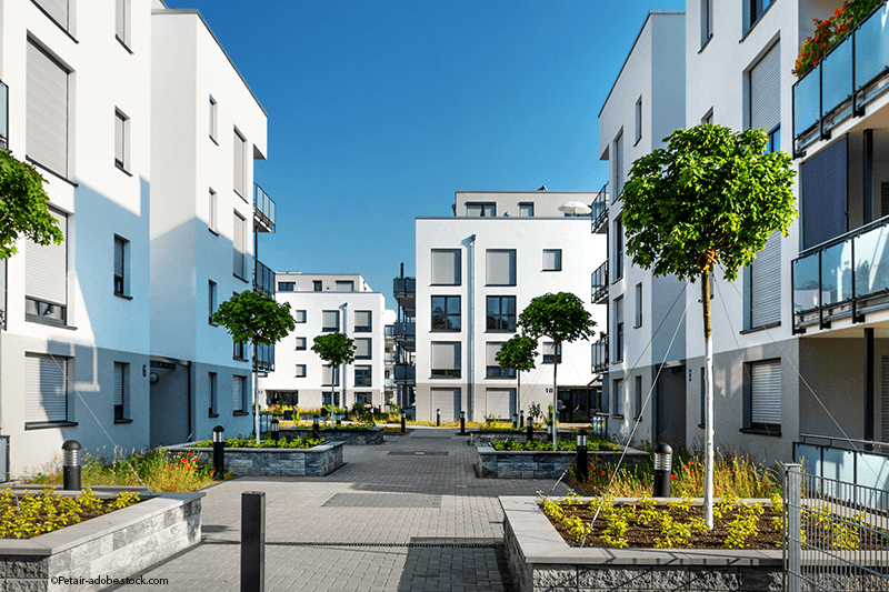Moderner Apartmentkomplex mit weißen Gebäuden, gepflasterten Wegen und gestutzten Bäumen unter klarem, blauem Himmel.