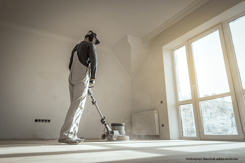 Eine Person in Schutzkleidung bedient eine Bodenschleifmaschine in einem hellen, sonnendurchfluteten Raum mit großen Fenstern.