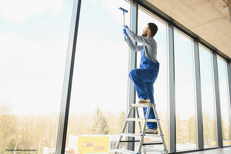Eine Person in blauem Arbeitsoverall steht auf einer Trittleiter und benutzt einen Abzieher, um ein hohes Glasfenster zu reinigen.