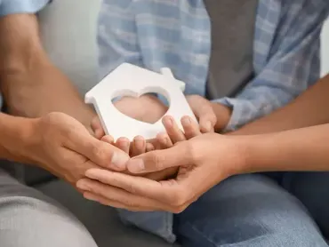 Una pareja sostiene una pequeña casa con un corazón en sus manos.