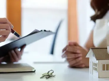 Una mujer está sentada en una mesa con un portapapeles y una maqueta de casa.