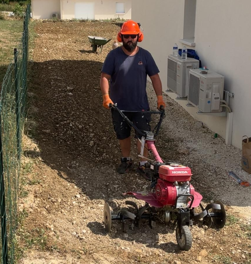 Un homme portant un équipement de sécurité utilise une motobineuse rouge sur une pente rocheuse à côté d'un bâtiment blanc.