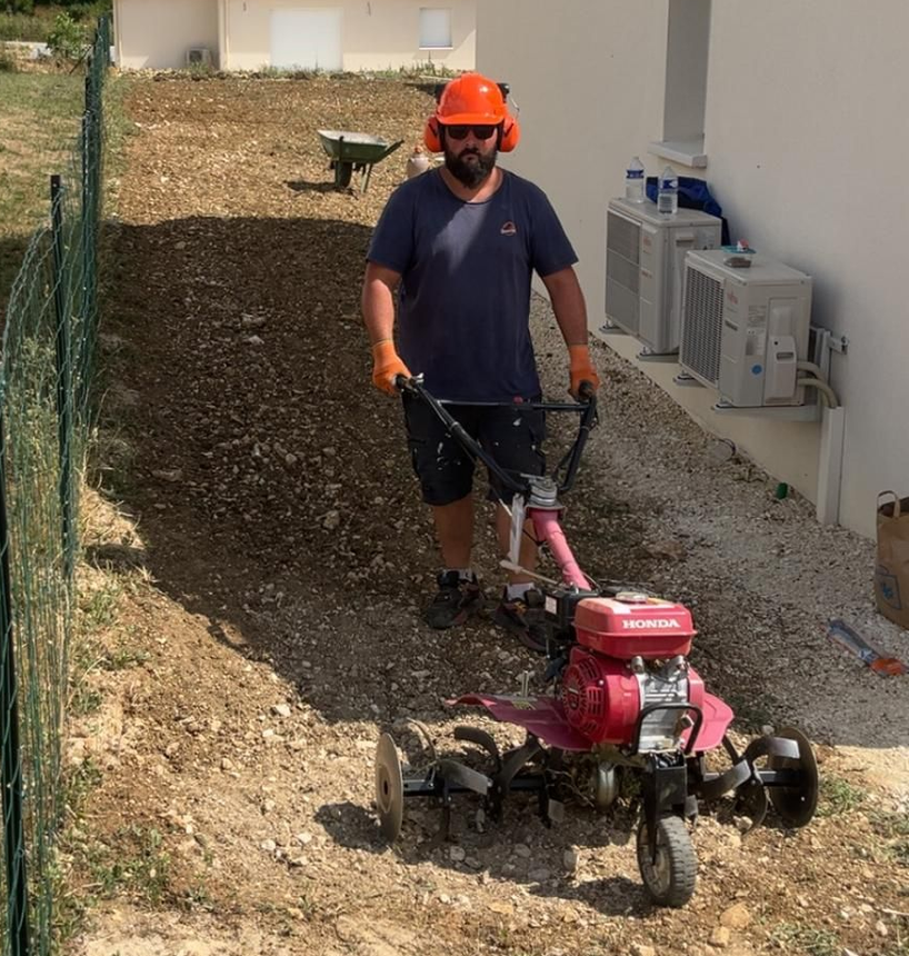 Un homme portant un équipement de sécurité utilise une motobineuse rouge sur une pente rocheuse à côté d'un bâtiment blanc.