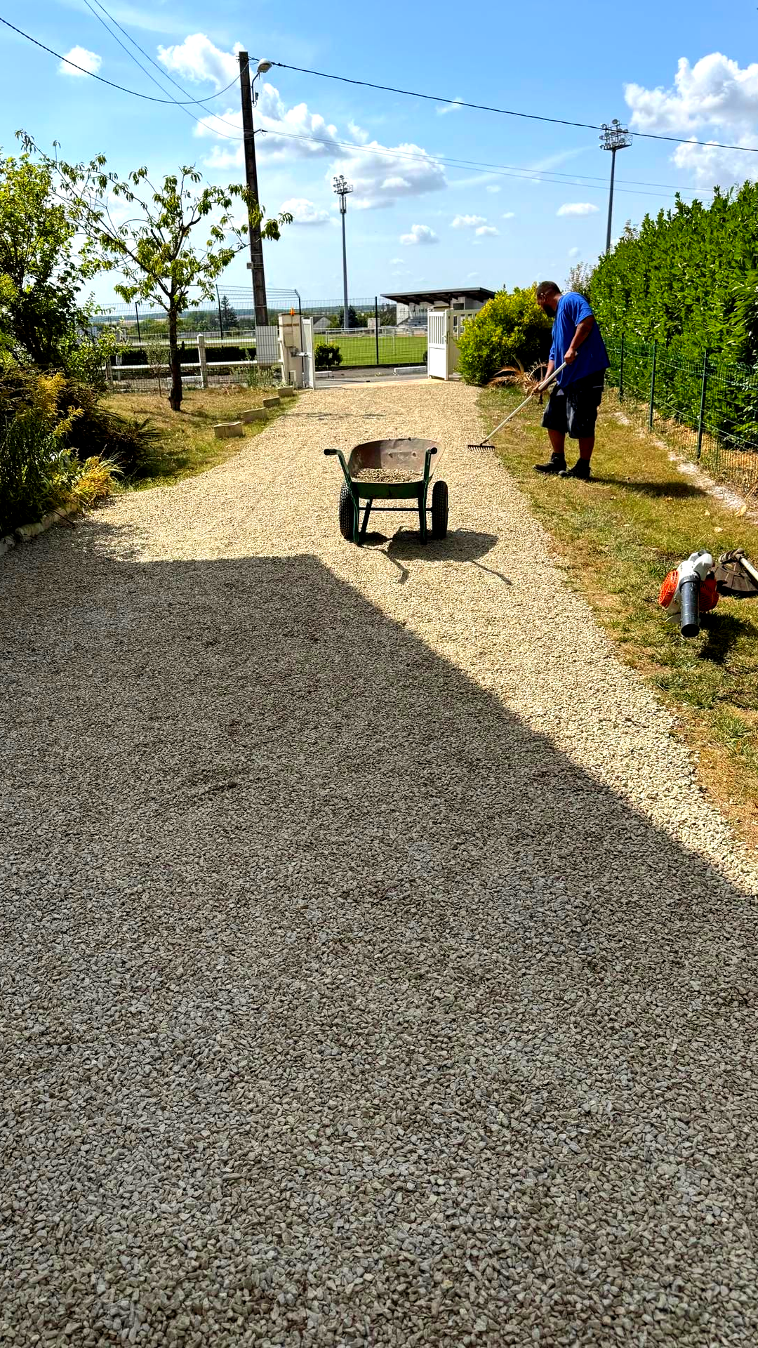 Allée de gravier avec une personne et une brouette ; le ciel est bleu et le décor est à l'extérieur.