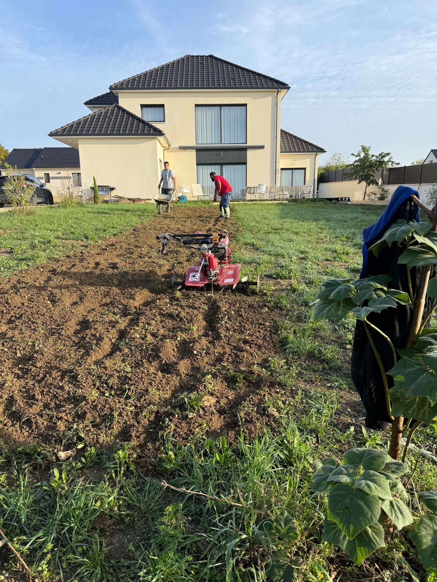 Deux personnes labourant la terre avec une machine dans une cour, une maison en arrière-plan.