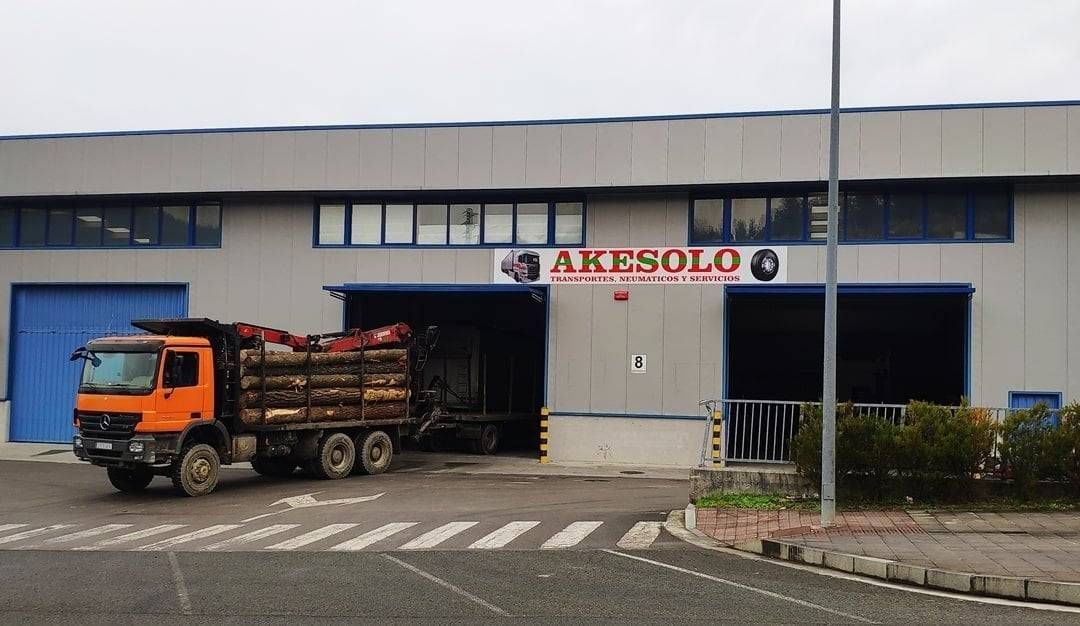 Un camión naranja descarga troncos en un almacén industrial azul con la etiqueta “AKRSELO”.