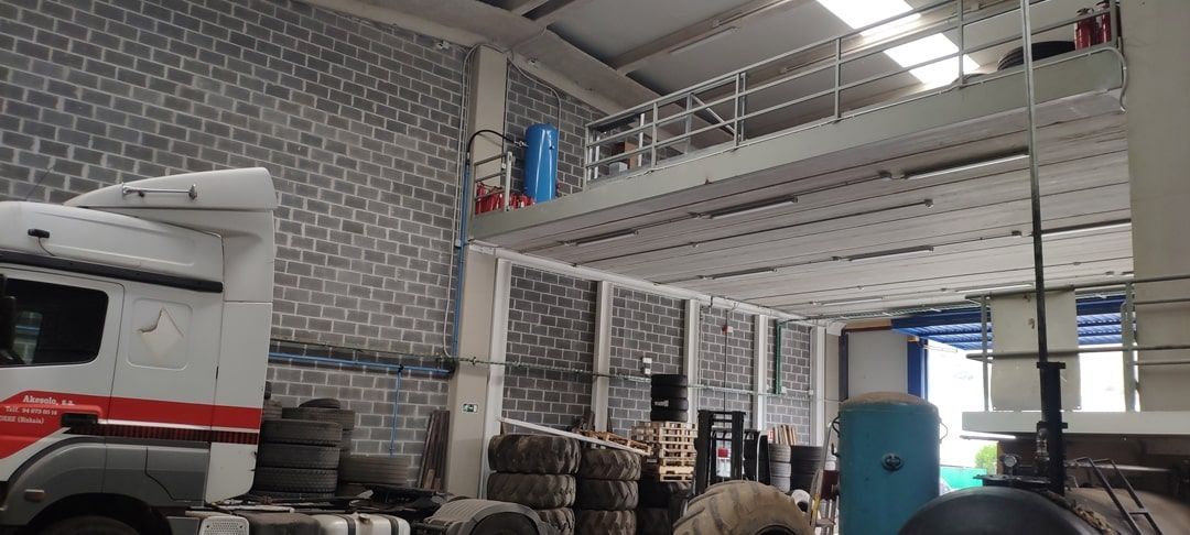 Taller mecánico interior con una furgoneta blanca, neumáticos apilados y un barril azul bajo un altillo metálico.