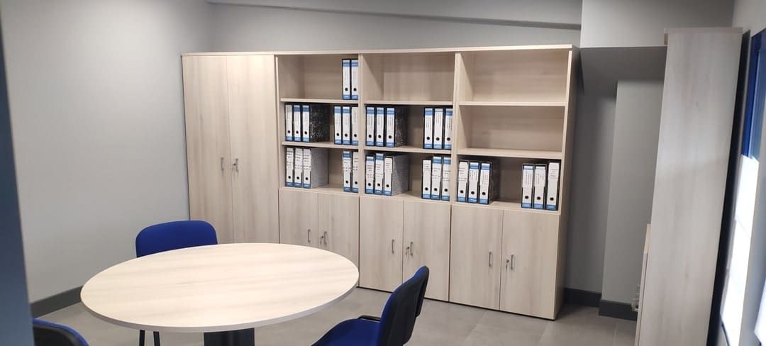 Sala de reuniones de oficina con mesa redonda, sillas azules y armarios de madera con carpetas.