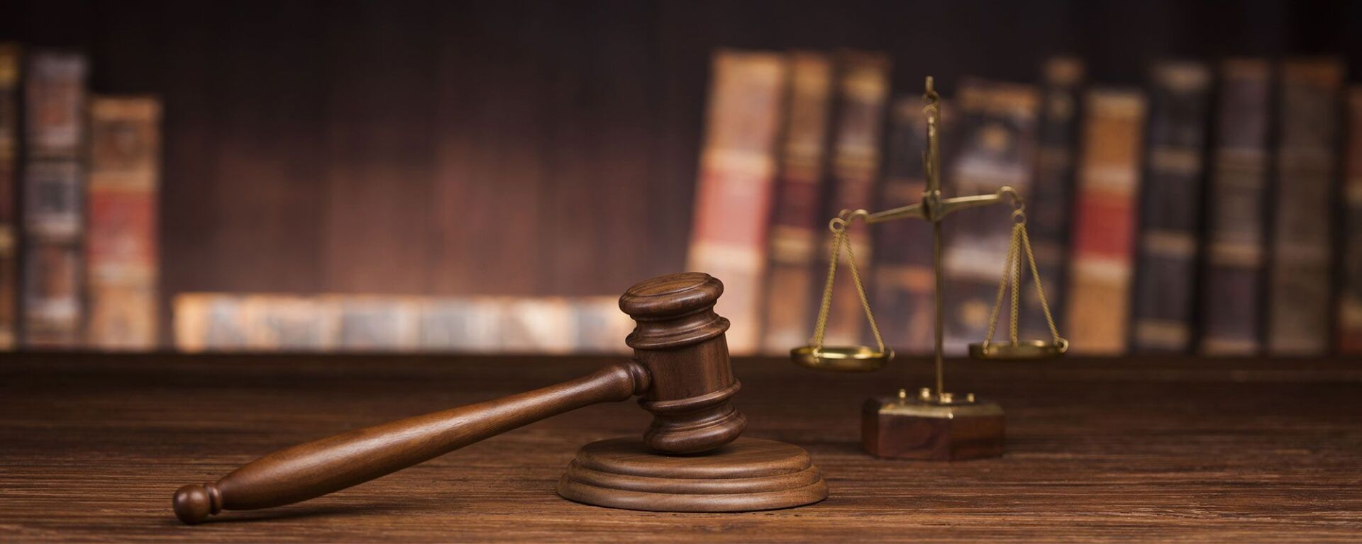 Ein Justitiahammer und eine Justitiawaage auf einem Holzschreibtisch, im Hintergrund ein Bücherregal.