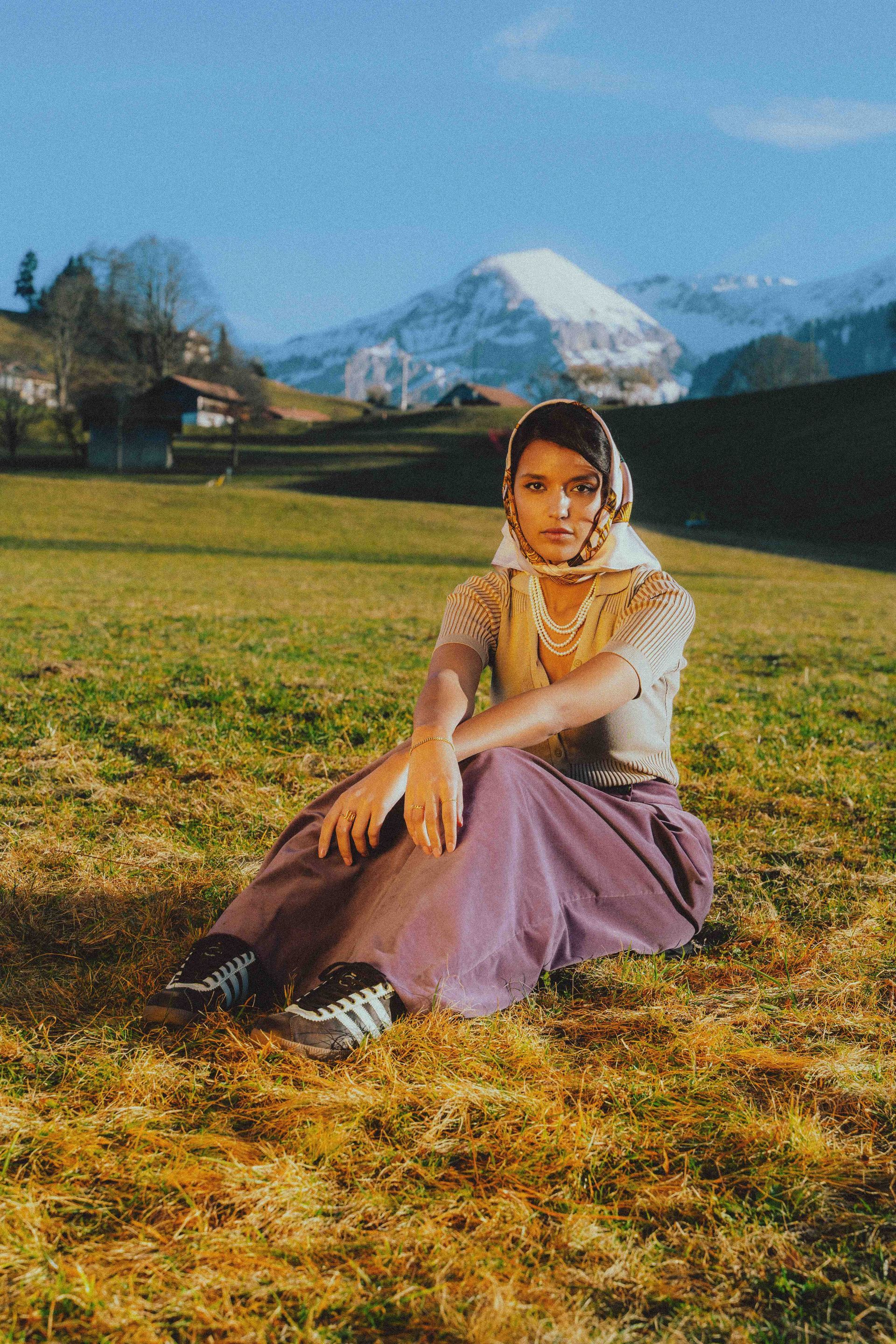 Eine Frau sitzt auf einem Feld im Gras mit Bergen im Hintergrund.