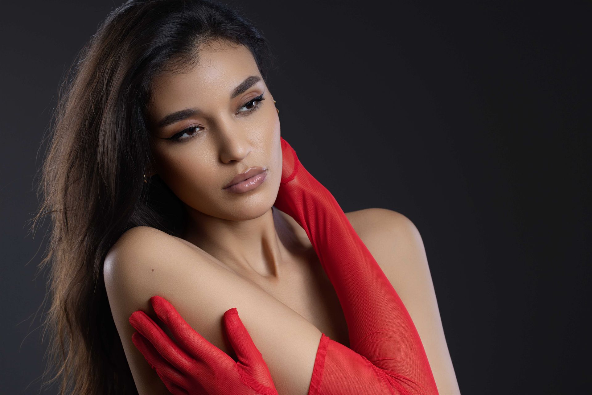 Frau mit langen dunklen Haaren, roten Handschuhen, blickt mit neutralem Gesichtsausdruck zur Seite. Schwarzer Hintergrund.