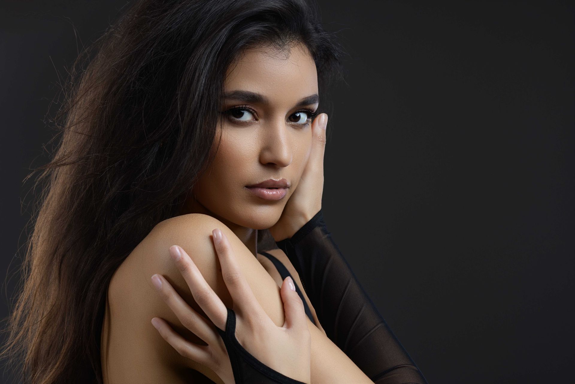 Frau mit dunklem Haar und Make-up, posiert vor einem grauen Hintergrund, Hand auf Gesicht und Schulter.