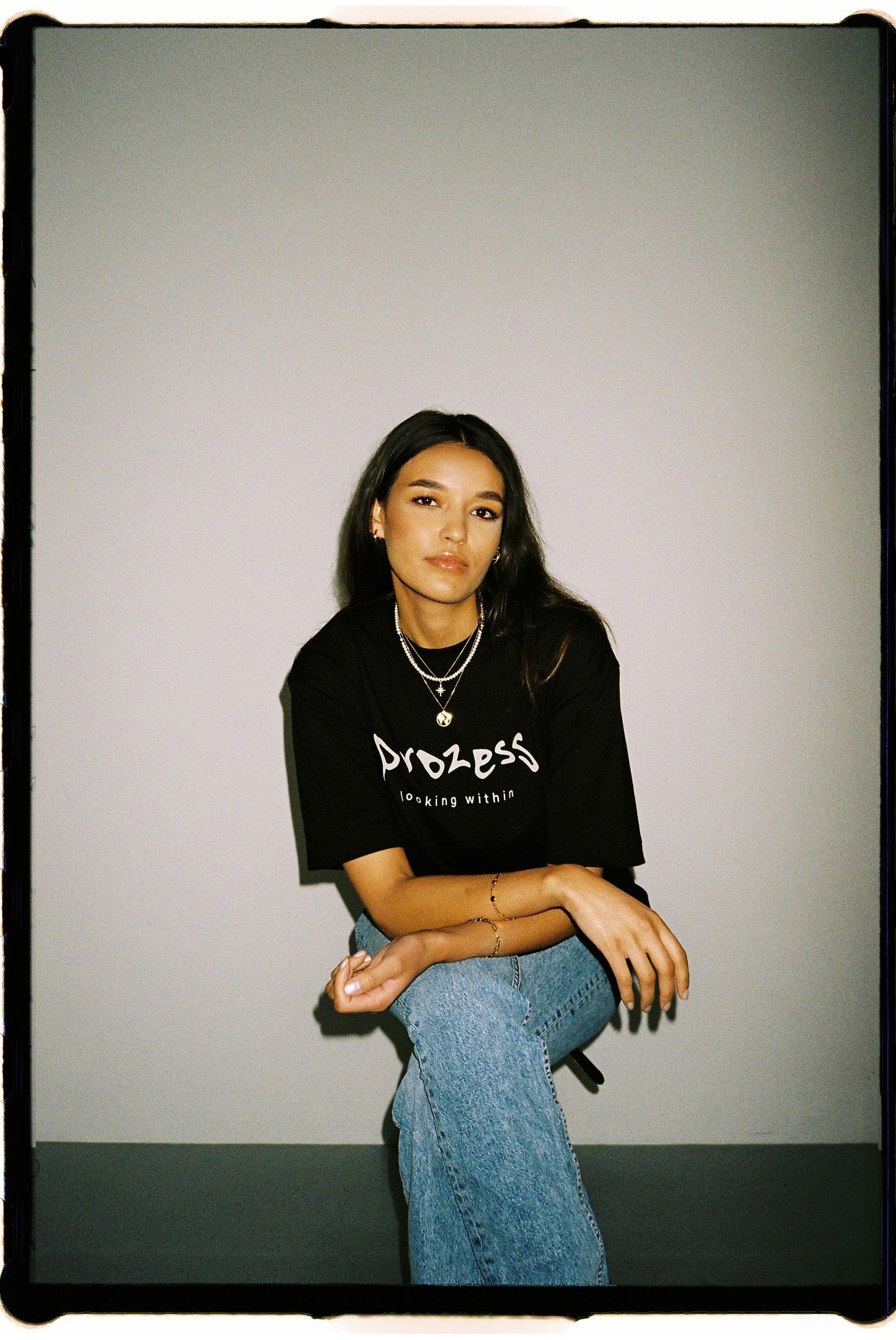 Eine Frau in schwarzem T-Shirt und Jeans sitzt vor einer Wand.