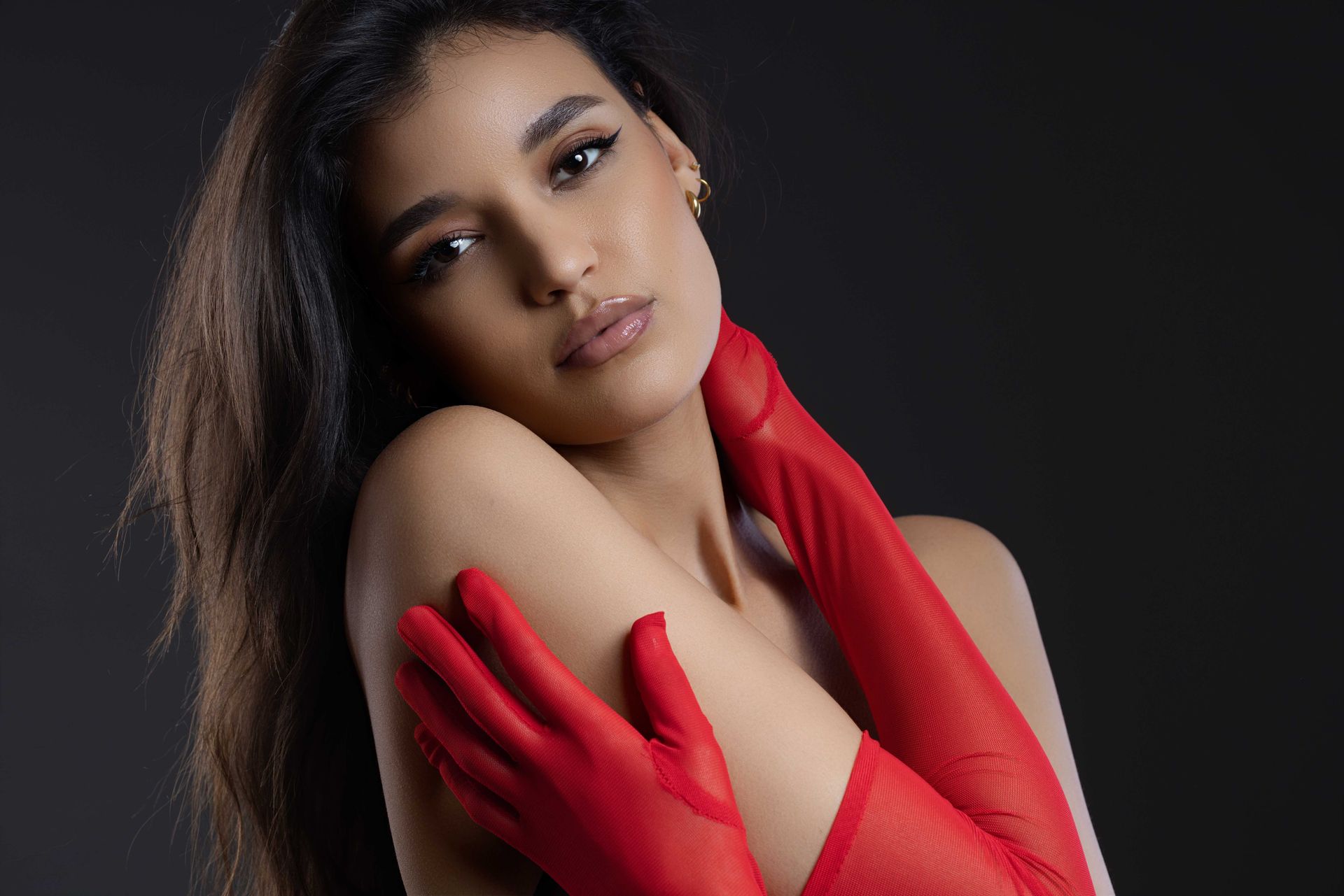 Frau mit langen dunklen Haaren und roten Handschuhen blickt in die Kamera. Vor einem dunklen Hintergrund legt sie die Arme um ihre Schultern.