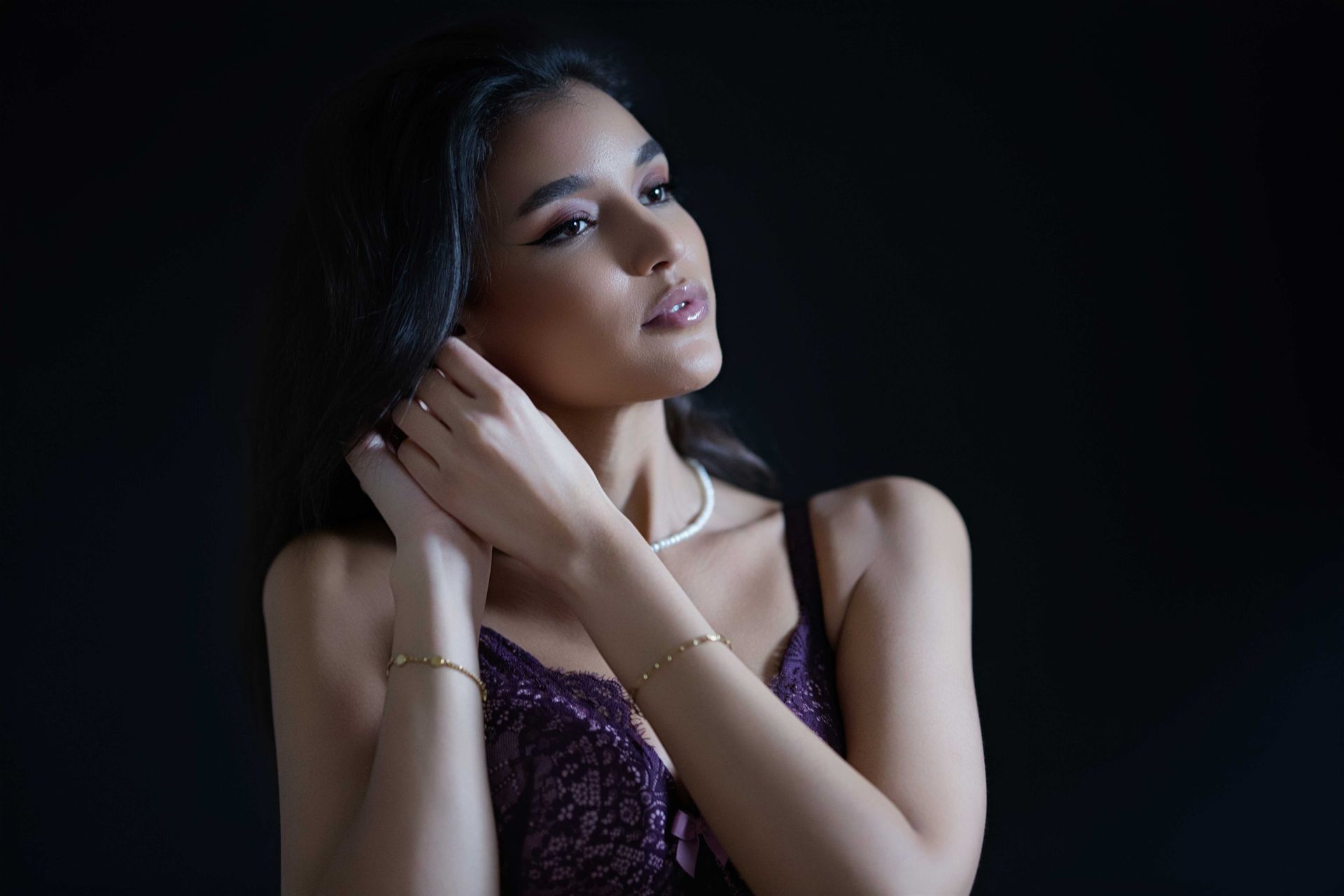 Frau in dunkelviolettem Spitzentop berührt ihr Ohr und trägt Schmuck vor schwarzem Hintergrund.