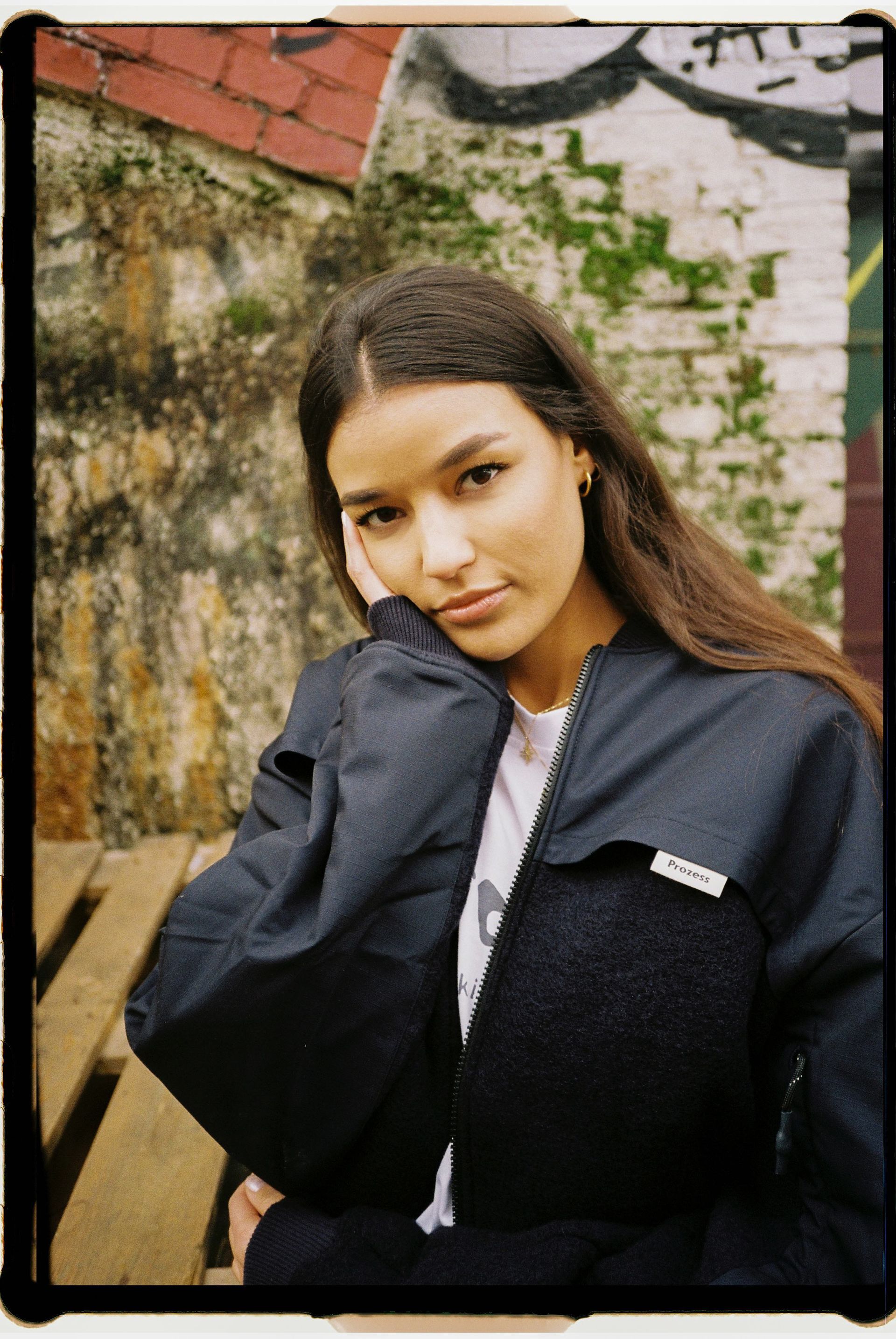 Eine Frau in einer schwarzen Jacke steht vor einer Backsteinmauer.