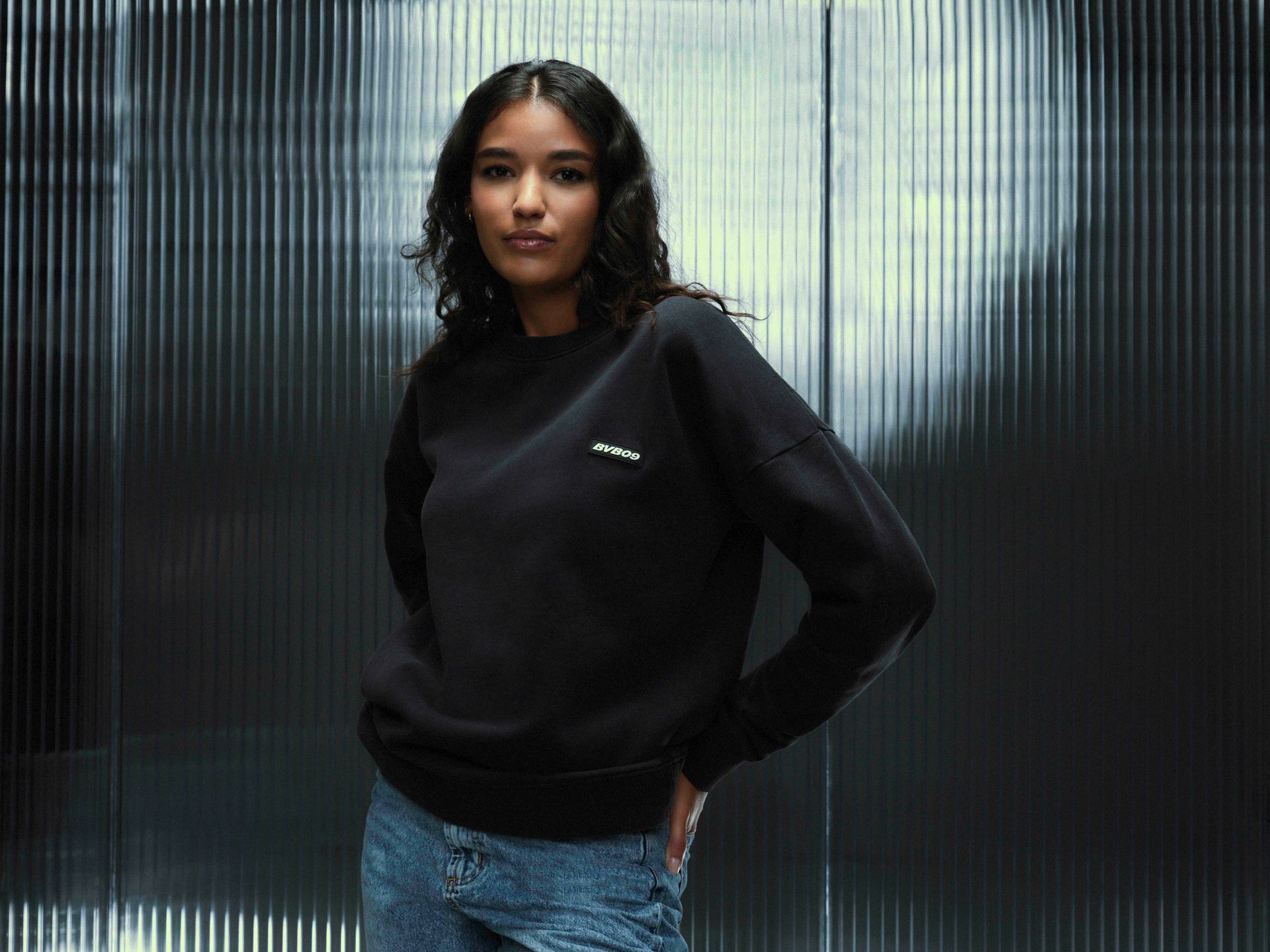 Frau in schwarzem Sweatshirt und Jeans steht vor einer strukturierten silbernen Wand.