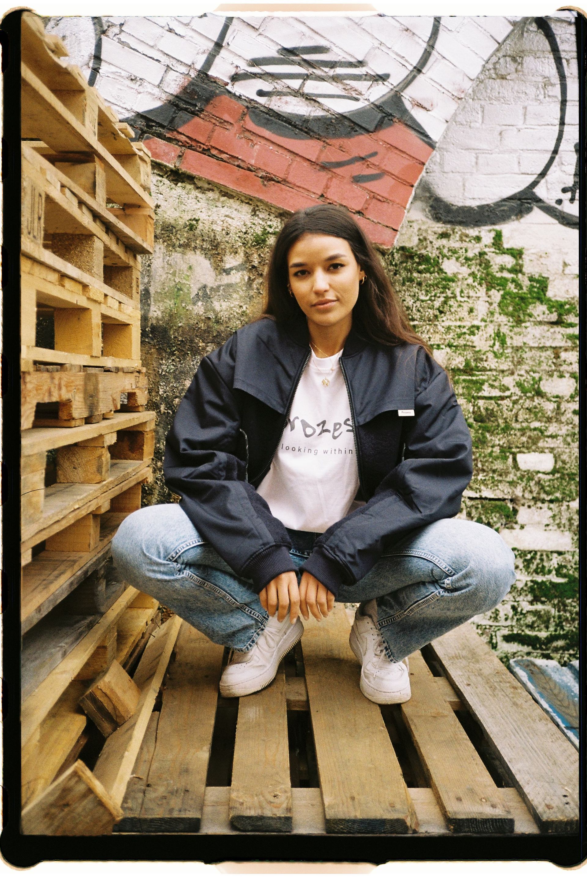 Eine Frau hockt auf einer Holzpalette vor einer Backsteinmauer.