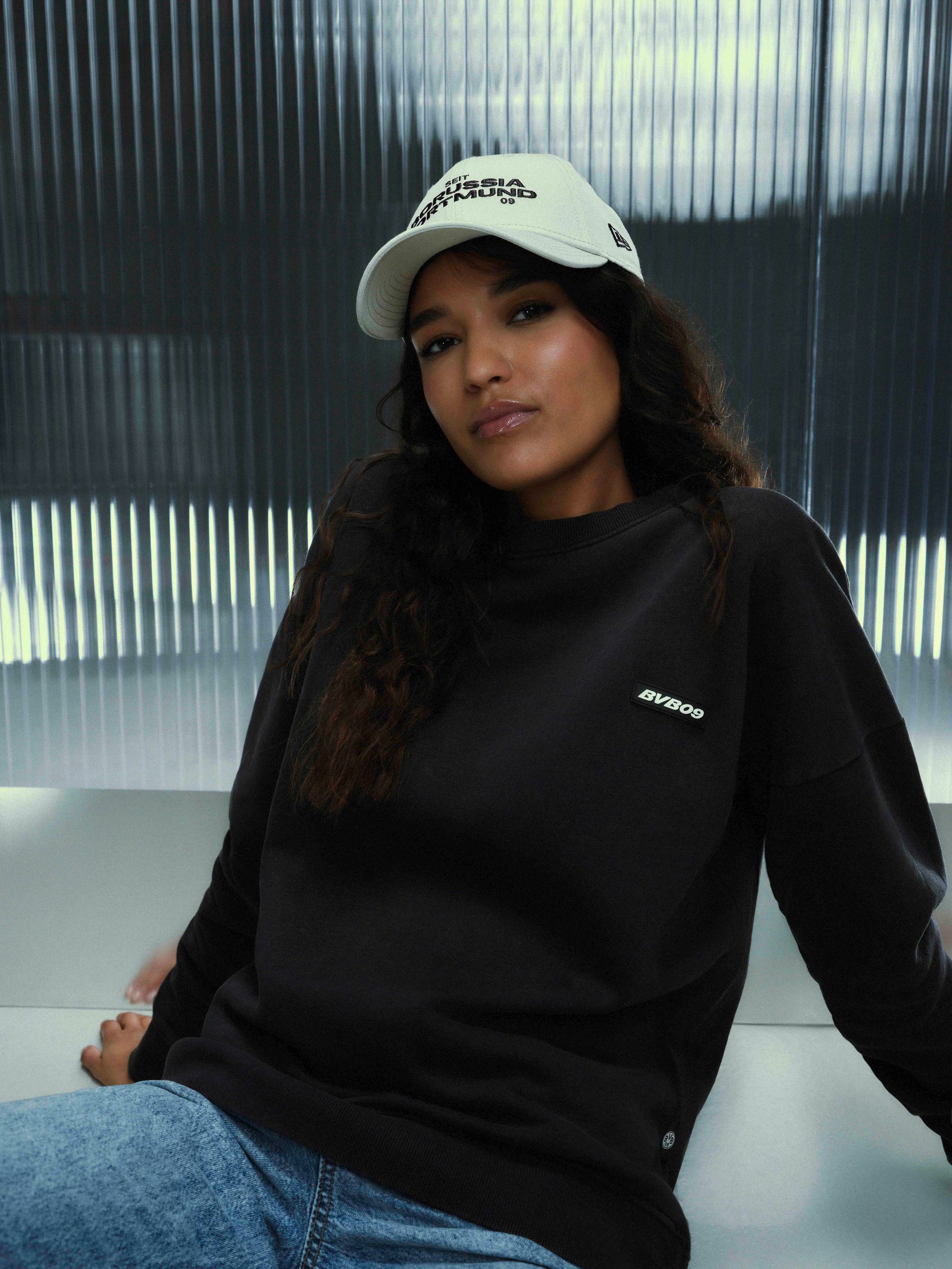 Eine Frau in einem schwarzen Sweatshirt und einer weißen Mütze sitzt lächelnd in hellblauen Jeans vor einem strukturierten grauen Hintergrund.