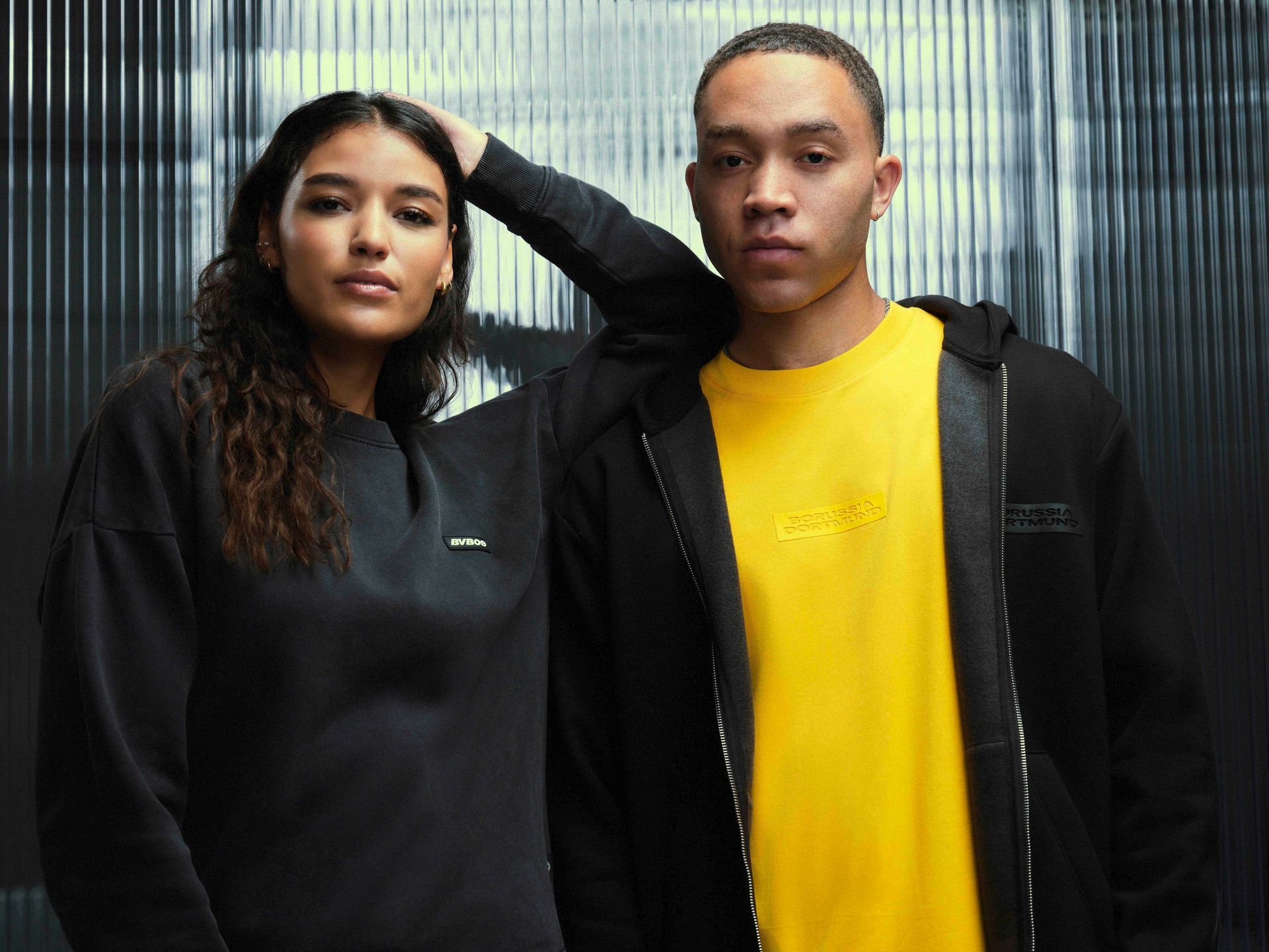 Mann im gelben Hemd und Frau im schwarzen Hemd posieren vor grauem Hintergrund.