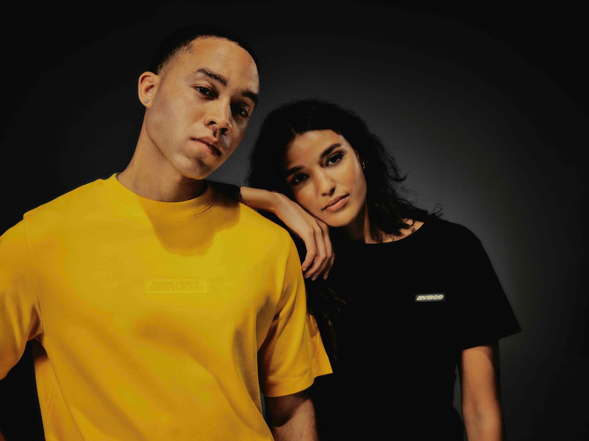 Mann im gelben Hemd und Frau im schwarzen Hemd posieren vor dunklem Hintergrund.