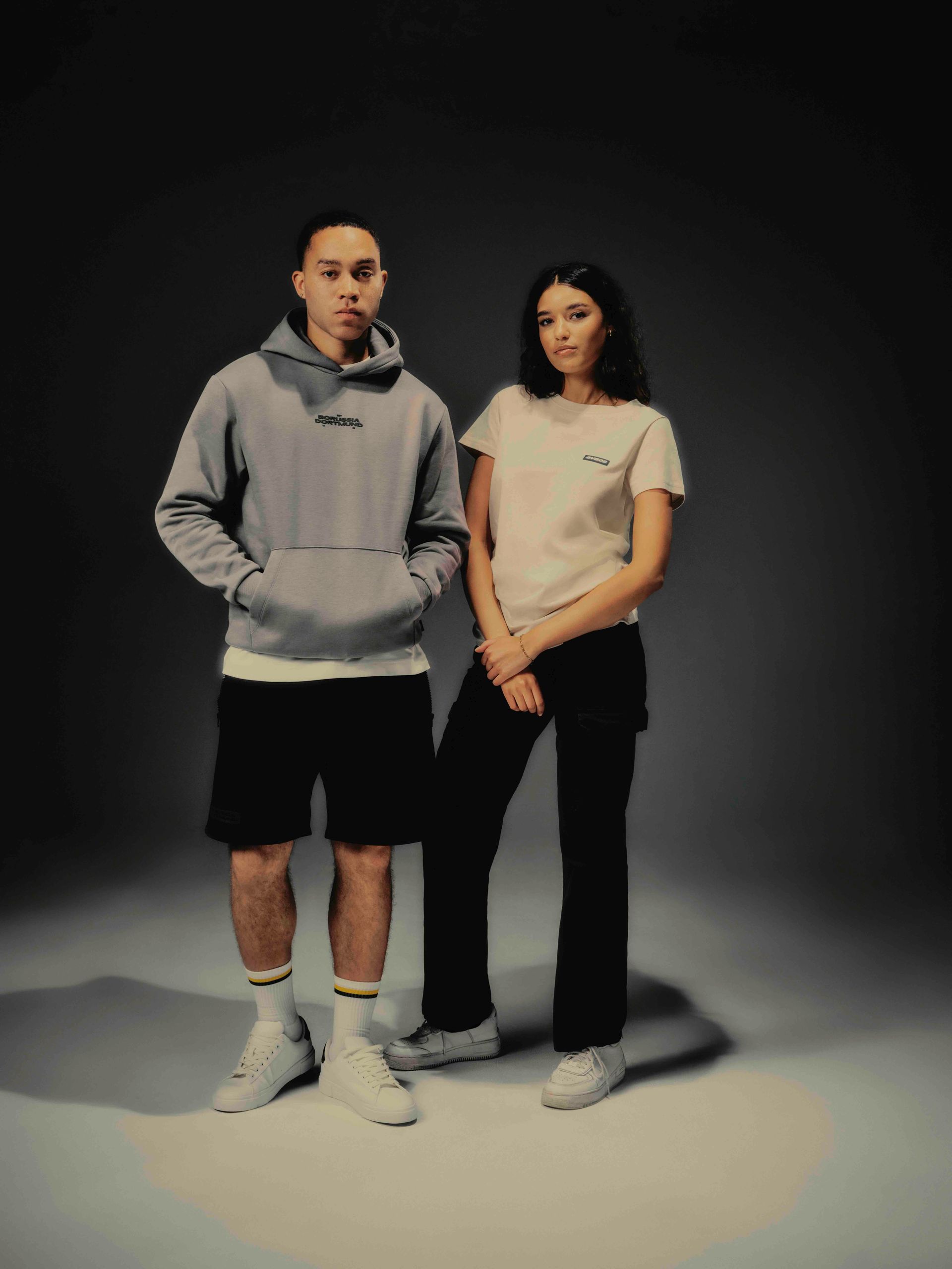 Mann in grauem Kapuzenpullover und Shorts, Frau in hellbraunem T-Shirt und schwarzer Hose, posiert. Dunkler Studiohintergrund.