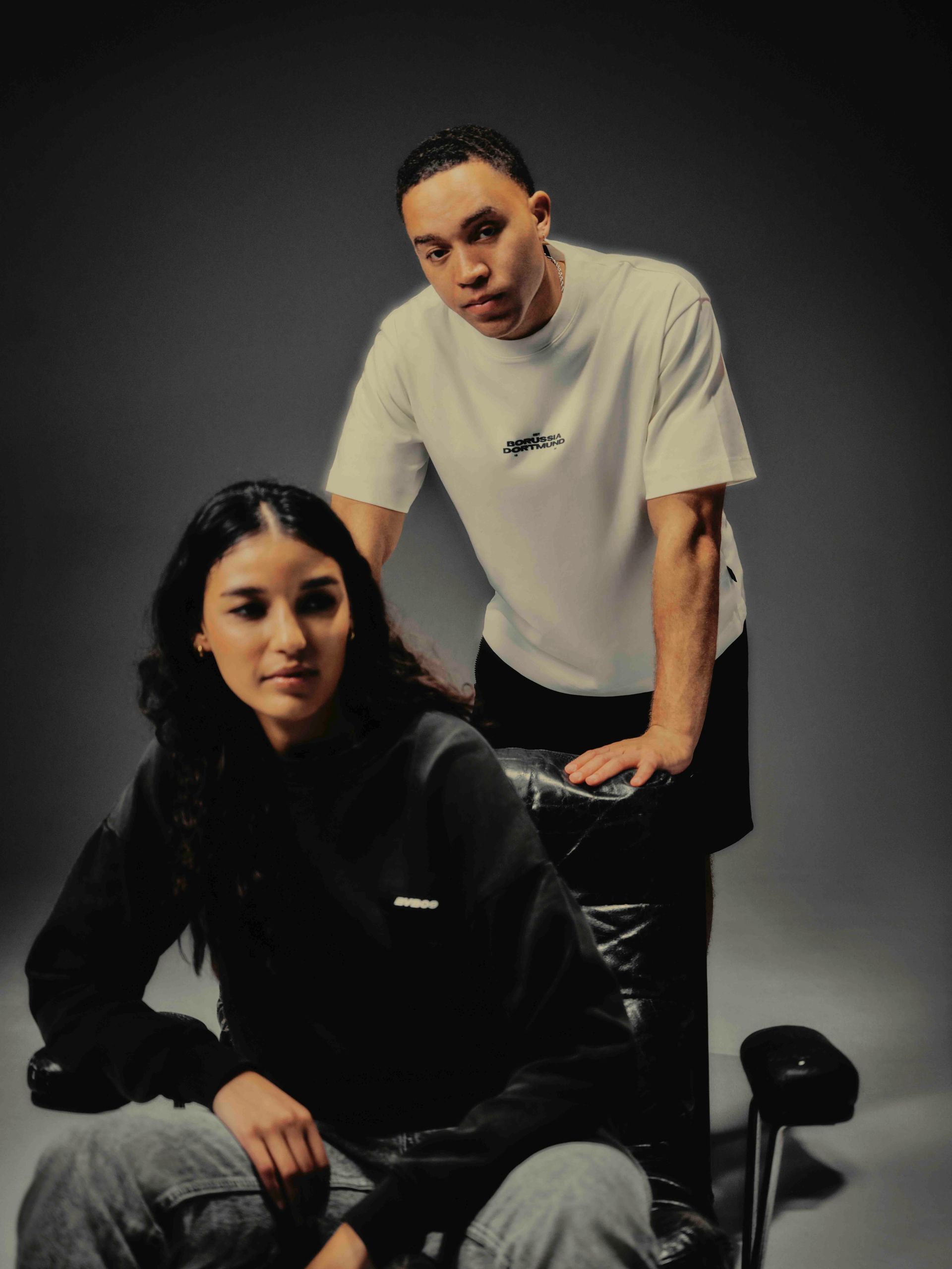 Eine Frau und ein Mann in einem Studio; die Frau sitzt auf einem Stuhl, der Mann steht hinter ihr, beide blicken in die Kamera.