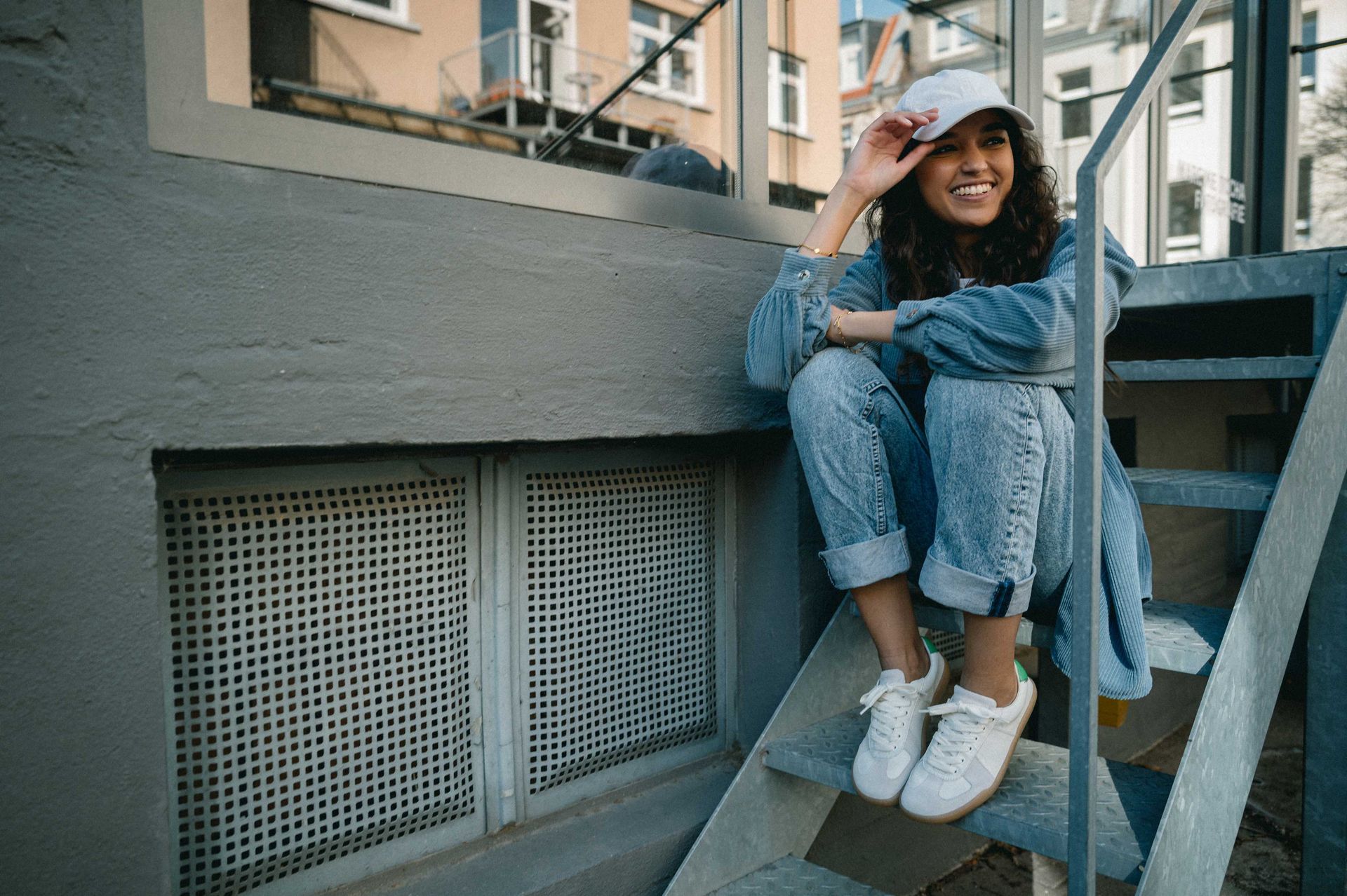 Frau in Jeanskleidung und weißer Mütze sitzt lächelnd auf Außentreppe. Graues Gebäude im Hintergrund.