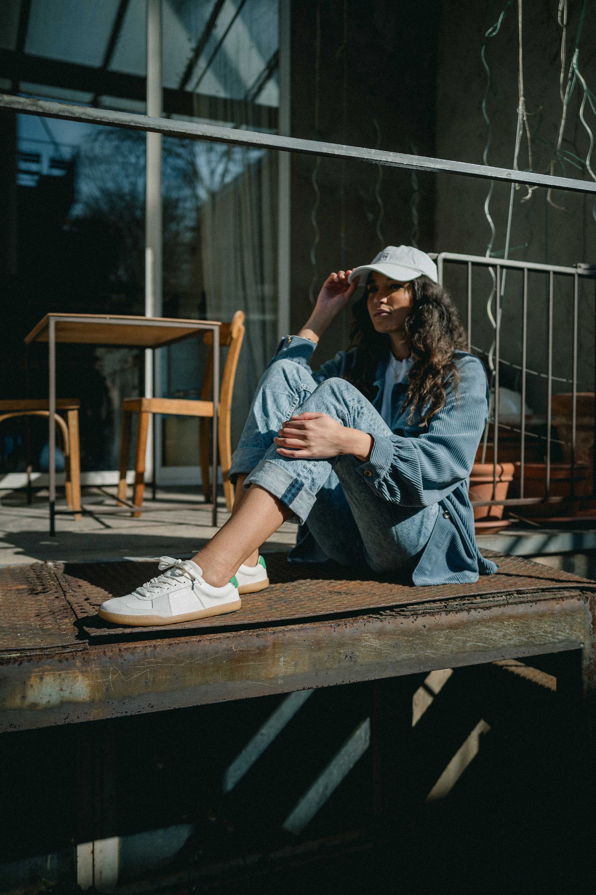 Eine Frau sitzt im Freien auf einer rostigen Metalloberfläche. Sie trägt eine Jeansjacke, Jeans und eine weiße Mütze und das Sonnenlicht wirft Schatten.