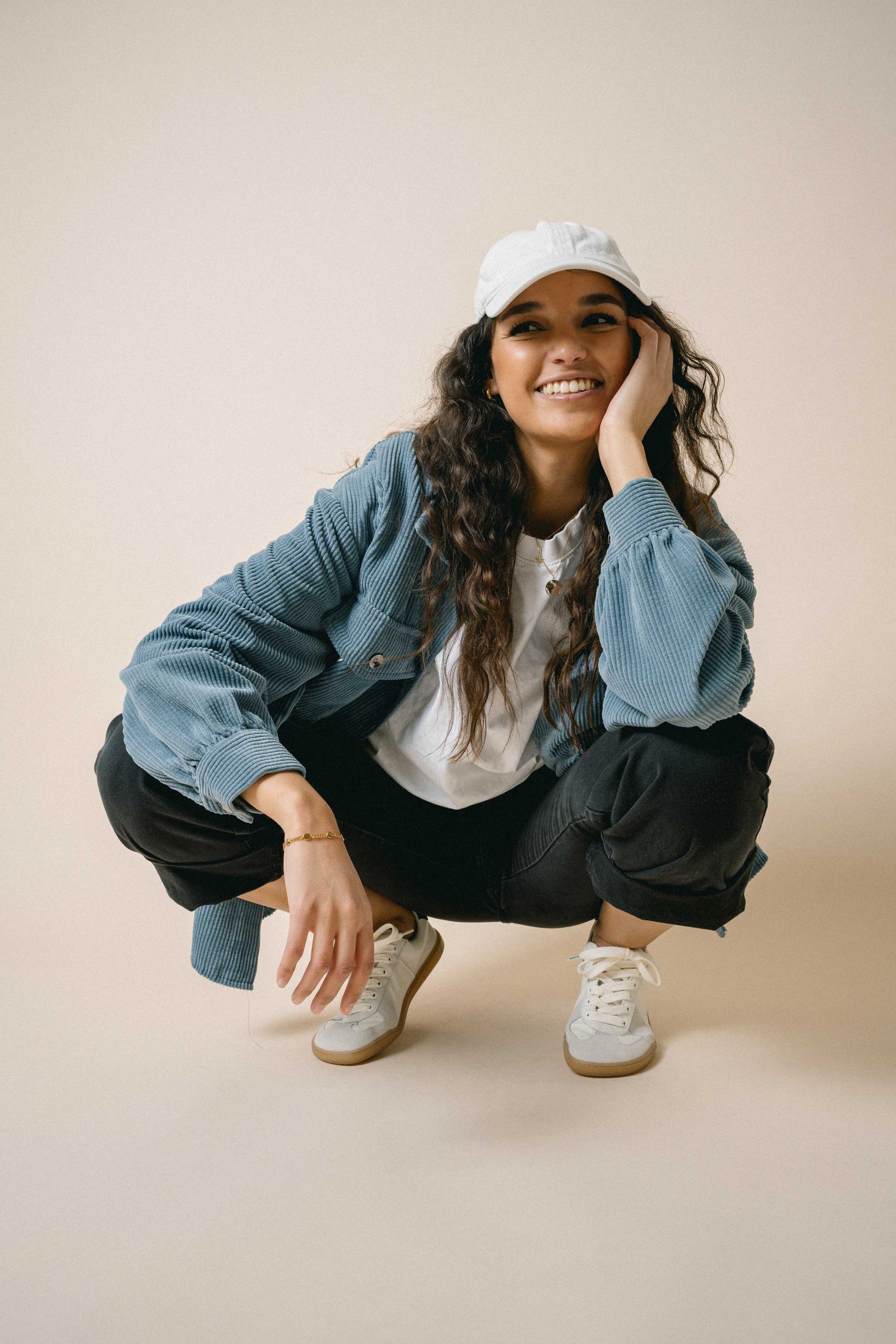 Frau mit lockigem braunem Haar, trägt eine weiße Mütze, eine blaue Jacke, eine schwarze Hose und Turnschuhe, hockt und lächelt. Studioeinstellung mit beigem Hintergrund.