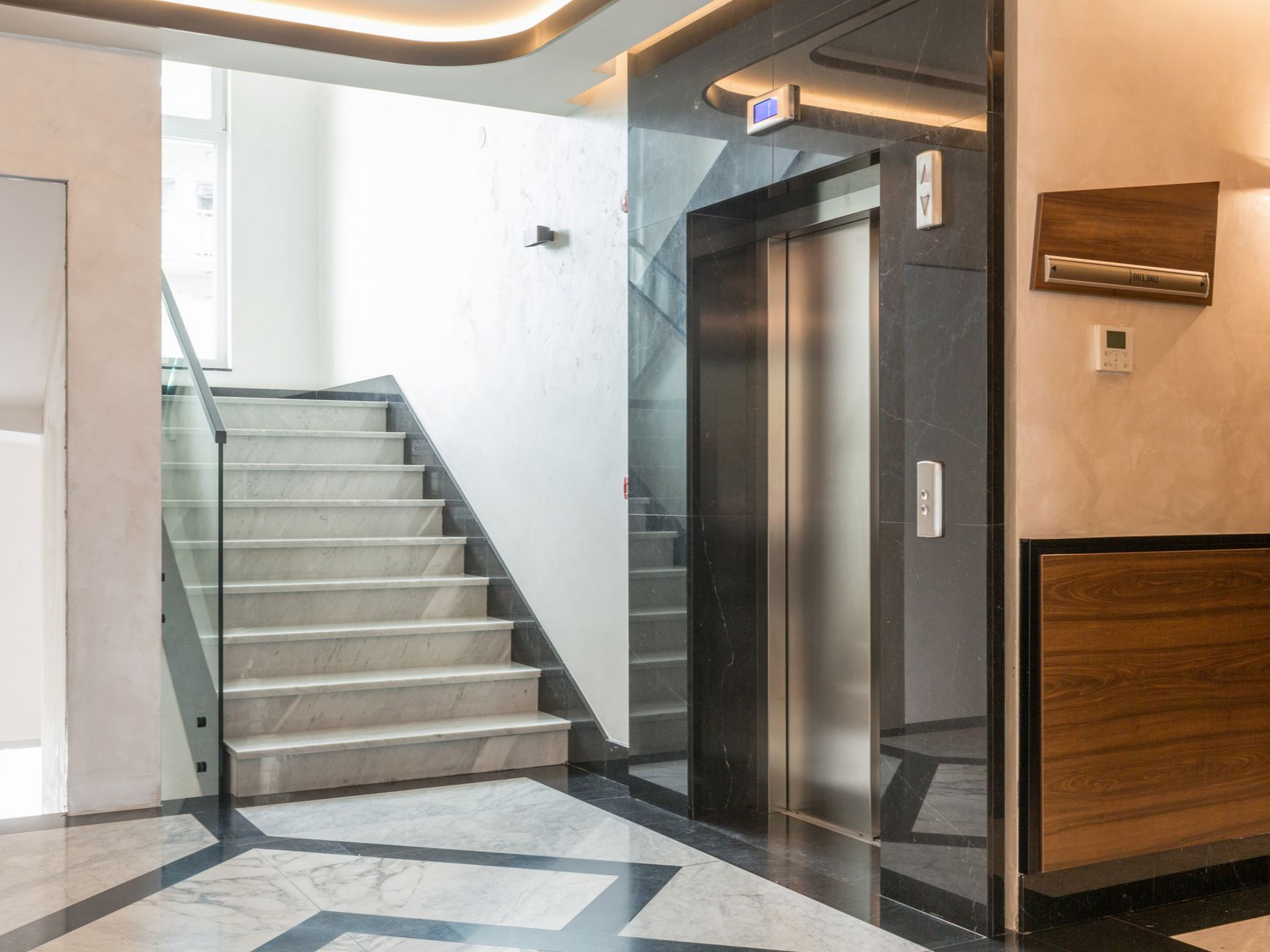Hall d'entrée moderne avec un escalier en marbre, une balustrade en verre et une porte d'ascenseur en acier inoxydable.
