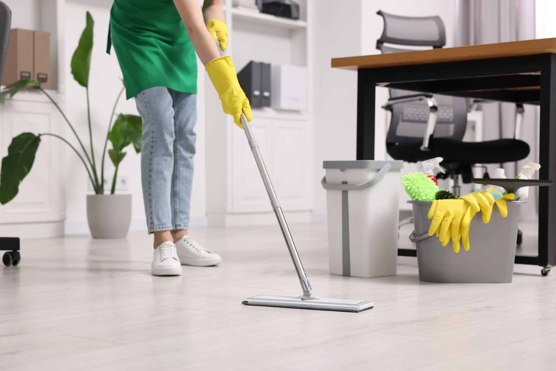 Une personne portant des gants en caoutchouc jaunes et un tablier vert lave le sol clair d'un bureau.