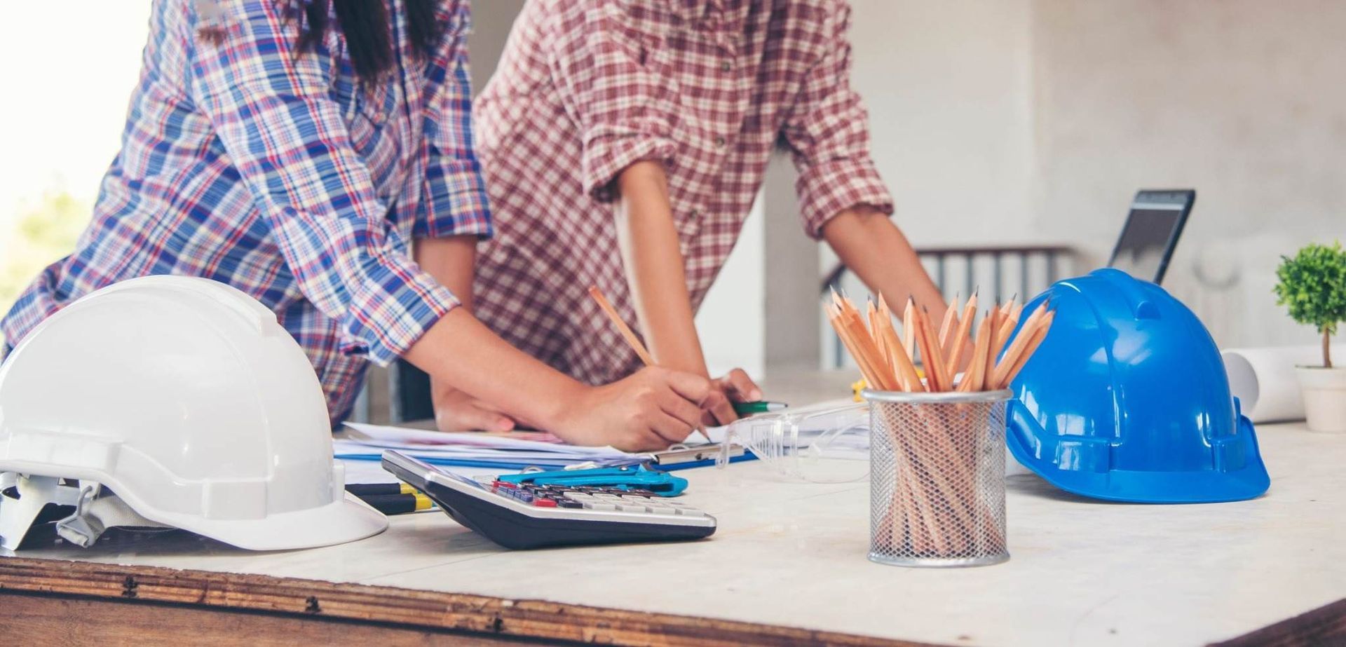 Un obrero de la construcción dibuja planos en una mesa con cascos blancos y azules y lápices cerca.