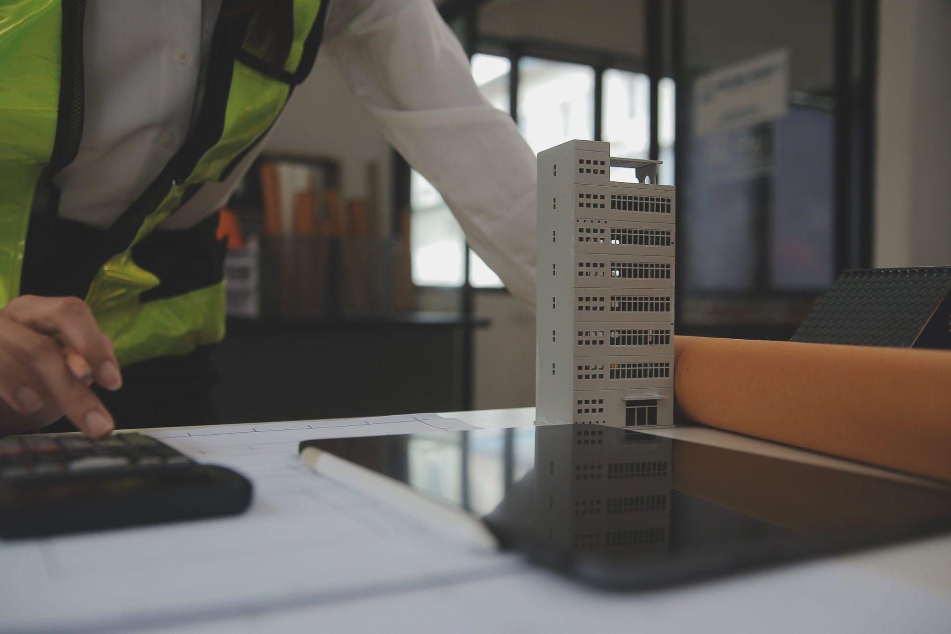 Escritorio de arquitecto con una maqueta de un edificio y una tableta, junto a un trabajador con un chaleco reflectante.