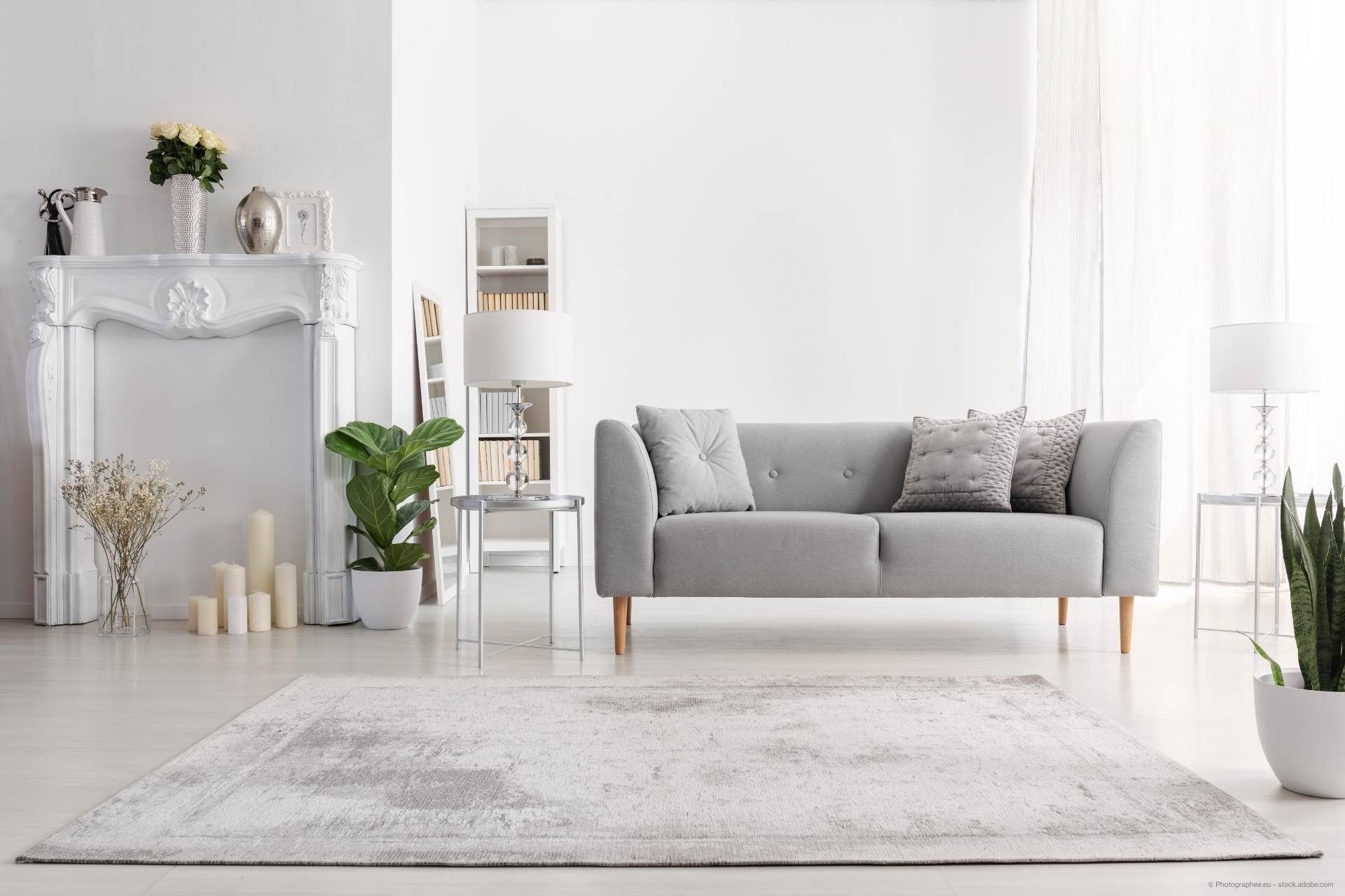 Ein stilvolles, minimalistisches Wohnzimmer mit einem grauen Sofa, elegantem Dekor, einem weißen Kamin und Grünpflanzen, alles in natürliches Licht getaucht.