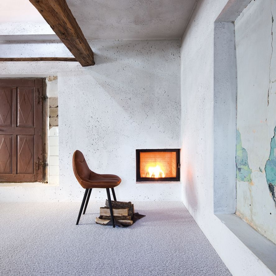 Ein kahler Raum mit Betonwänden und einem hellen Teppichboden, auf dem ein Stuhl und Brennholz für den Kamin liegt