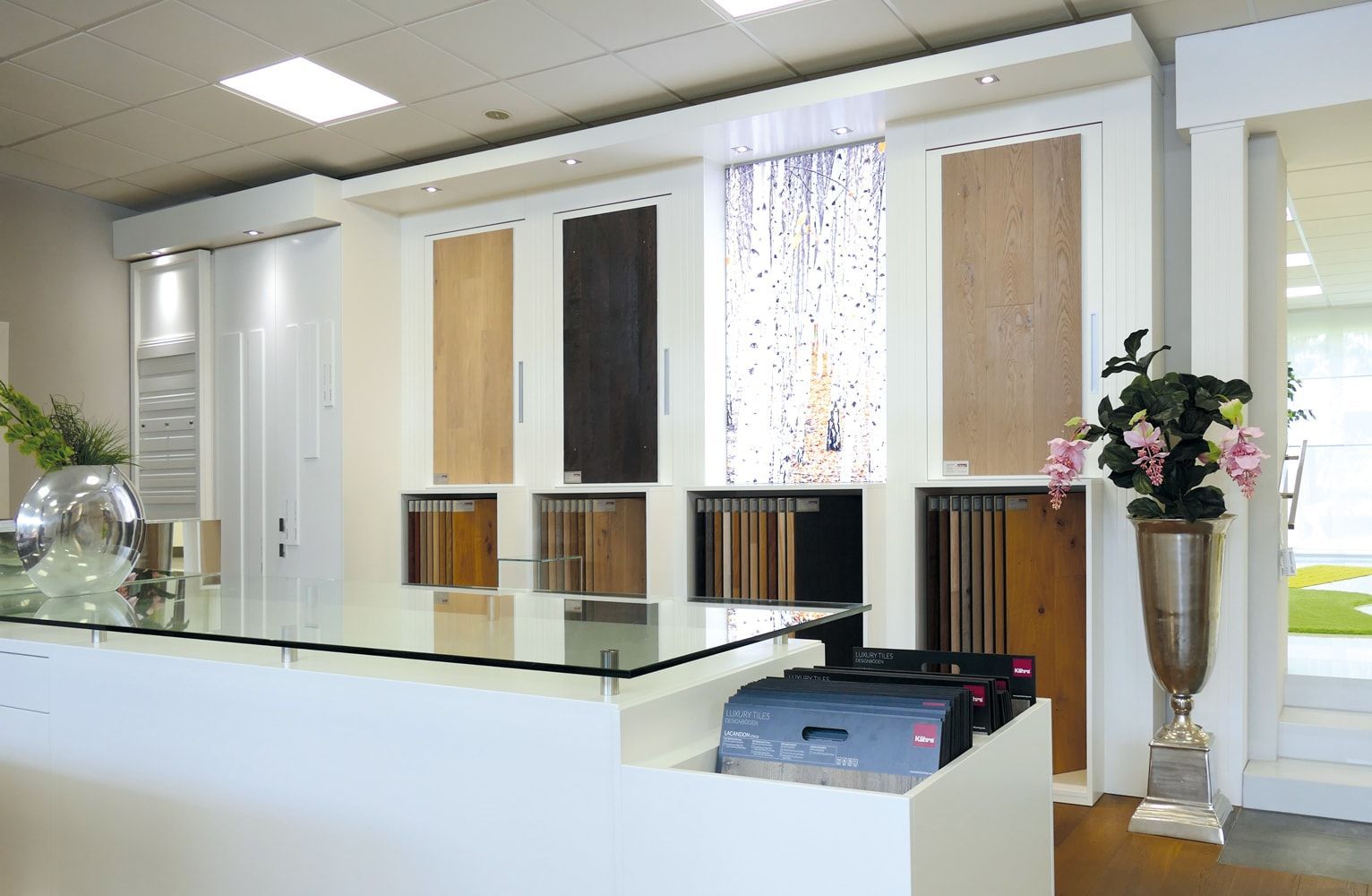 Ein moderner Ausstellungsraum mit einem Glastisch, Holzpaneel-Mustern und dekorativen Pflanzen in eleganten Töpfen.