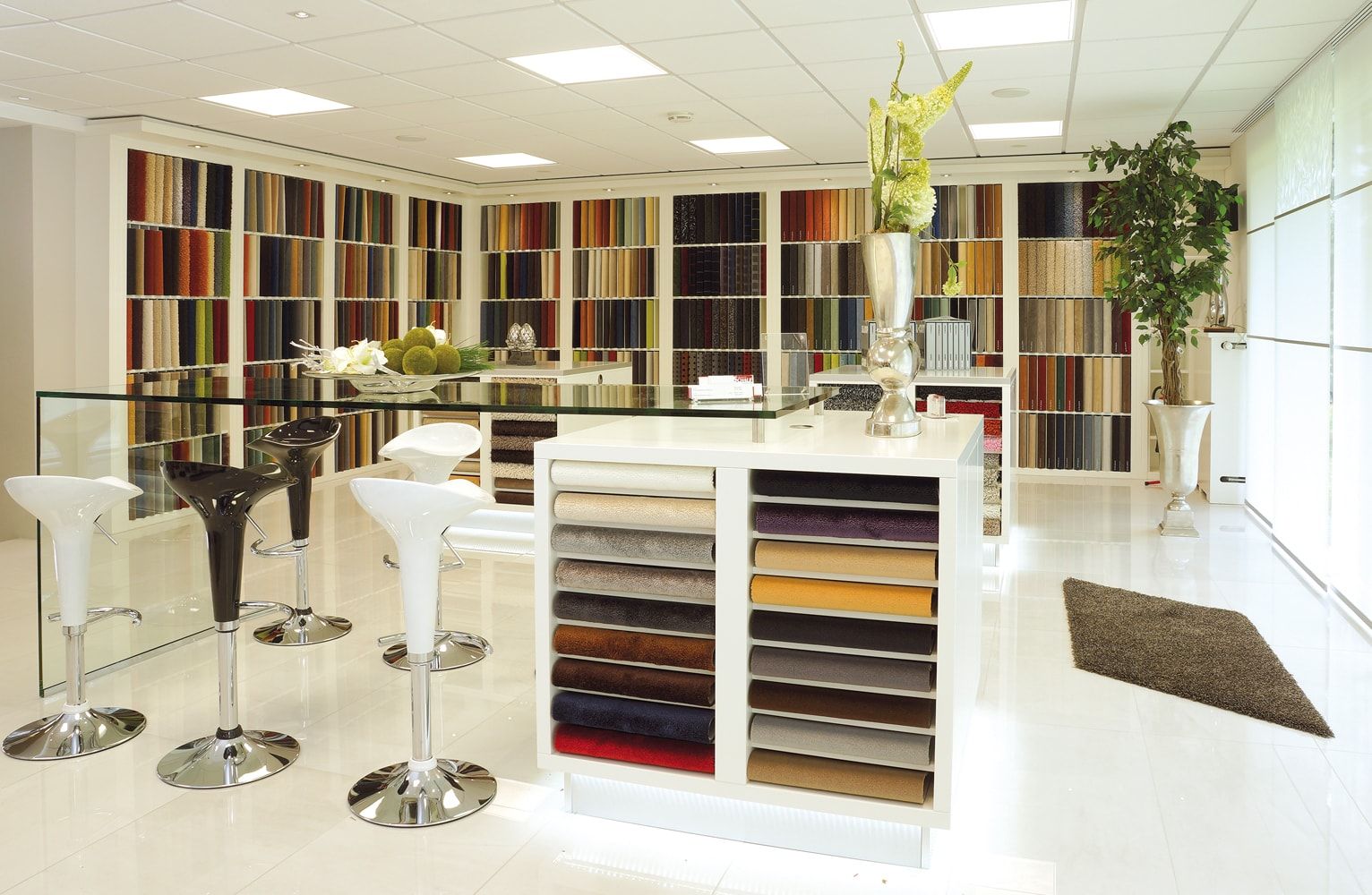 Stilvolles Interieur eines Stoffladens mit einem Glastisch, modernen Hockern und bunten Stoffrollen in Regalen.