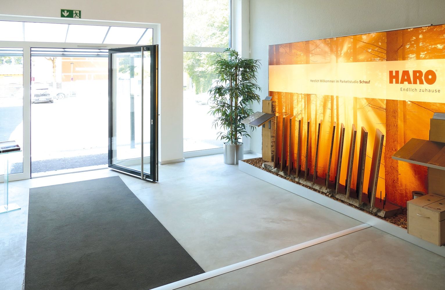 Heller Eingang eines Bodenbelagsstudios mit Holzmustern, einem Willkommensschild und einer Pflanze neben der offenen Tür.