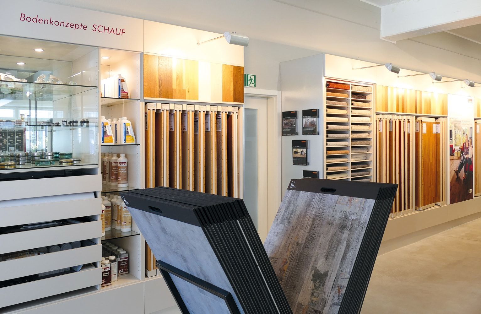Innenraum eines Ausstellungsraums für Bodenbeläge mit verschiedenen Holz- und Laminatmustern, Reinigungsprodukten und Schautafeln.