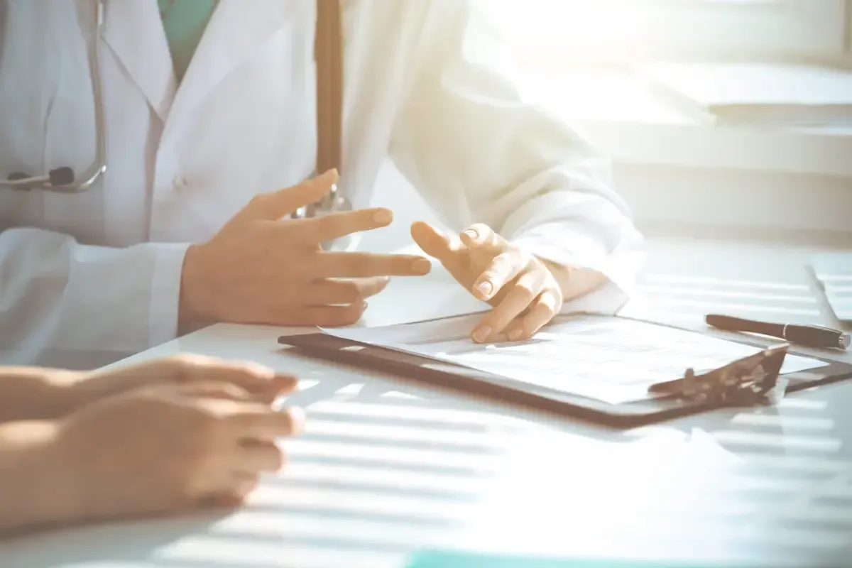 Médico con bata blanca hace un gesto al paciente con un portapapeles bajo la luz del sol.