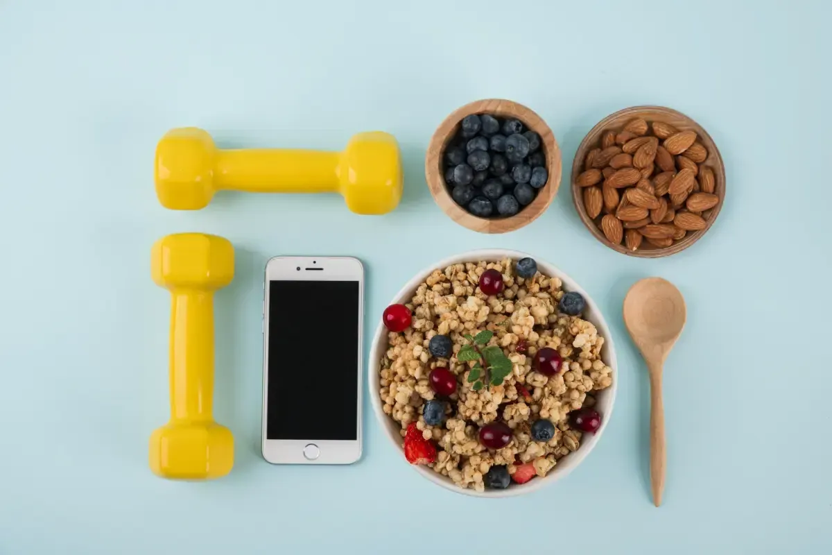 Mancuernas amarillas, teléfono inteligente, tazón de granola, fruta, nueces y cuchara sobre una superficie azul.