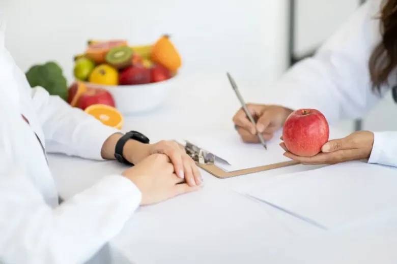 Una dietista le muestra una manzana a un paciente y toma notas. Un frutero está sobre la mesa.