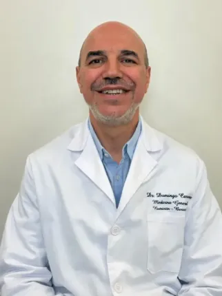 Médico sonriente con bata blanca y camisa azul, frente a una pared blanca.