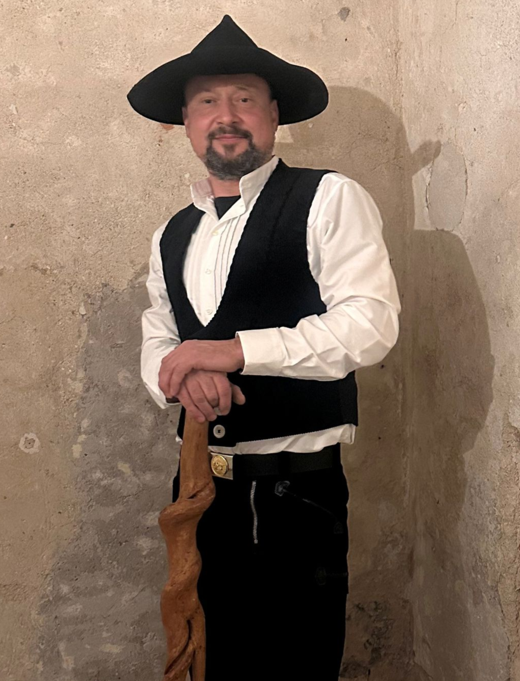 Ein Mann mit schwarzem Hut und schwarzer Weste lehnt sich an einen geschnitzten Holzstab. Weißes Hemd, schwarze Hose, schlichter Wandhintergrund.