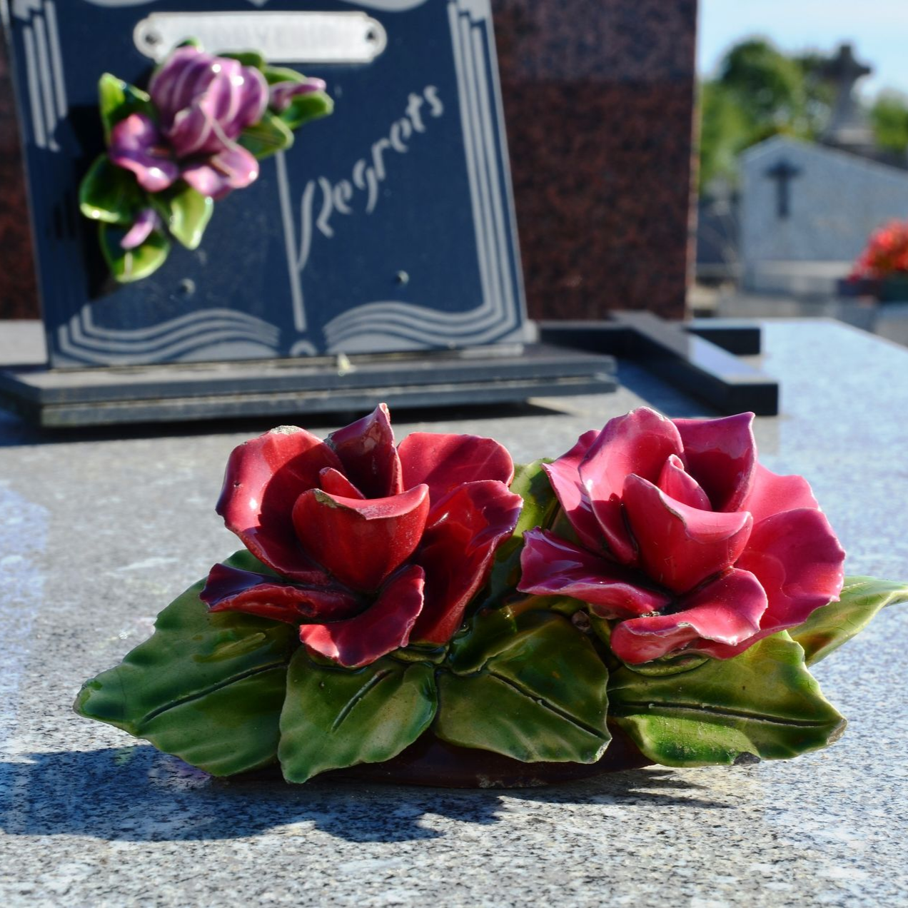 Roses en céramique rouges sur une pierre tombale grise, avec un mémorial sombre en arrière-plan.